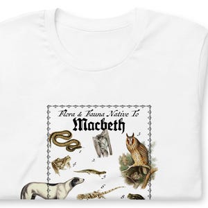 May include: A white t-shirt featuring a graphic print titled Flora & Fauna Native To Macbeth. The design includes numbered illustrations of a snake, bat, owl, toad, lizard, and other creatures arranged within a decorative black border frame.