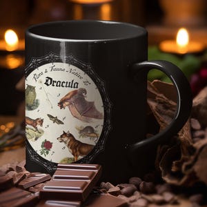 Op de afbeelding: Een zwarte keramische mok met een cirkelvormige afbeelding met de tekst Flora & Fauna Native To Dracula. De illustratie bevat genummerde afbeeldingen van een uil, een vleermuis, een mot, een vos, een roos, een wolf en een muis in een rand.