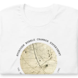 May include: A white t-shirt featuring a circular graphic of a hydrogen bubble chamber experiment. The text reads HYDROGEN BUBBLE CHAMBER EXPERIMENT and 1981 in a black sans-serif font. The central image shows thin, dark, intersecting particle tracks.