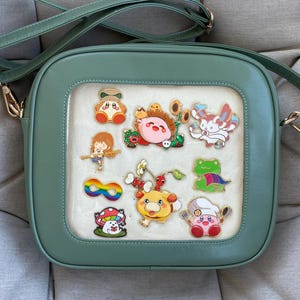 May include: A sage green crossbody bag with a clear window displaying colorful character pins. The bag has a square shape, a long adjustable strap, and gold-colored hardware. The pins feature various cartoon characters in bright colors.