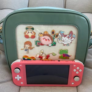 May include: A pale green shoulder bag with a clear window showcasing colourful character pins, and a coral pink Nintendo Switch Lite console. The bag has a gold chain strap. The pins include a rainbow, cherries, and a frog.