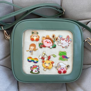 May include: A sage green crossbody bag with a clear front panel displaying colourful cartoon character pins. The bag has a square shape with rounded corners, a long adjustable strap, and gold-coloured hardware.