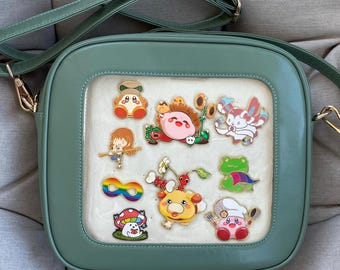 Könnte beinhalten: Eine salbeigrüne Umhängetasche mit einem transparenten Frontpanel, das bunte Cartoon-Charakter-Anstecknadeln zeigt. Die Tasche hat eine quadratische Form mit abgerundeten Ecken, einen langen, verstellbaren Riemen und goldfarbene Beschläge.