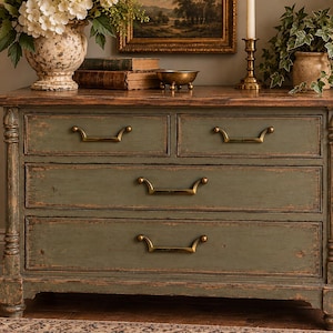 May include: A wooden dresser featuring a distressed sage green finish with visible wood grain and wear along the edges. It has two smaller top drawers and two larger bottom drawers, all equipped with curved brass-toned metal handles.