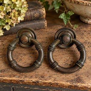 May include: Two circular metal drawer pulls with a dark, weathered bronze finish. Each pull features a round mounting plate and a hinged ring handle with decorative ribbed detailing along the circumference, showing a textured, aged patina.