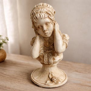 May include: A cream-colored bust sculpture of a woman with her hands near her face. The sculpture has detailed features, including a decorative headband, a pendant, and a floral design on the base. The bust is set on a wooden surface.