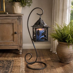 May include: A black metal lantern with a leaf-patterned frame hangs from a curved, scroll-shaped stand. The lantern contains a single blue pillar candle. The structure features a square base and a hook at the top for suspending the lantern.