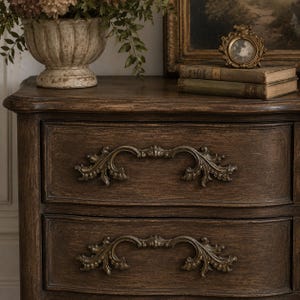 May include: A dark brown wooden dresser with two drawers, ornate brass handles, and a distressed finish. The top surface features a vase of flowers, books, and a framed picture, creating a vintage aesthetic.