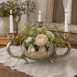 May include: A silver centerpiece with a floral arrangement and lit candles. The centerpiece has a bowl-like base with a petal design, and curved arms holding candle holders. The arrangement includes a pink flower and greenery.
