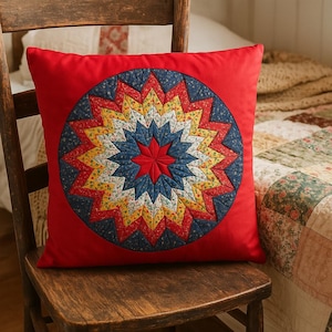 May include: A square red pillow with a circular starburst design. The starburst features a red center, surrounded by blue, yellow, and floral patterned segments. The pillow is placed on a wooden chair.