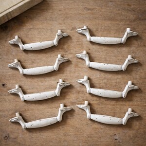 May include: Seven white, distressed-finish cabinet pulls arranged on a wooden surface. Each pull has a curved handle and decorative ends. The pulls are a creamy white color, with visible wear and tear.