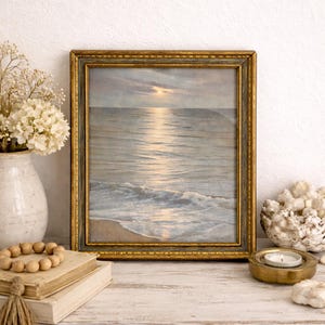 May include: A framed seascape painting with a gold ornate frame. The artwork depicts a sunset over the ocean, with the sun reflecting on the water. The painting is displayed on a white surface with decorative items.