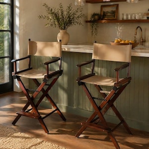 May include: Two brown wooden director's chairs with tan canvas seats and backs. The chairs have armrests and are positioned in front of a kitchen counter. The background includes a vase with greenery and a window.