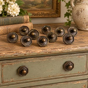 May include: A collection of ten octagonal cabinet knobs made of dark, weathered bronze metal. Each knob features a central circular protrusion and a flat, multi-sided base. Two of these knobs are installed on a green wooden drawer front.