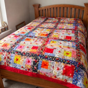 May include: A colorful patchwork quilt on a wooden bed frame. The quilt features squares of various colors, including red, yellow, blue, and pink, with floral accents. The bed frame is made of light-colored wood.