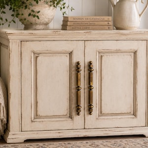 May include: A distressed off-white cabinet with two doors and brass-colored handles. The cabinet has a slightly weathered appearance, with visible wear and tear. A decorative vase, books, and a pitcher sit on top. A woven basket and a blanket are visible on the left.