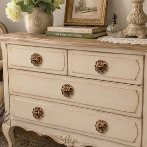 May include: Cream-colored chest of drawers with four drawers and ornate bronze-colored knobs. The chest has a distressed finish and a light-colored wooden top. The style is reminiscent of French country furniture.