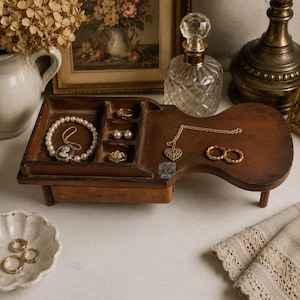 May include: A dark brown wooden jewelry organizer with a partitioned tray and a flat extended surface. The tray holds a pearl bracelet with a cameo, two small rings, and stud earrings. A gold heart necklace and hoop earrings rest on the flat area.