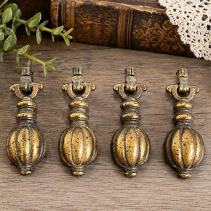 May include: Four antique brass drawer pulls with a gold finish. Each pull has a decorative, ribbed, oval-shaped body and a detailed top with a hinged handle. The pulls are arranged in a row on a wooden surface.