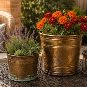 May include: Two hammered copper planters, one with lavender and sage, the other with orange and red marigolds. A decorative lantern sits nearby on a wrought iron table. The planters are outdoors, suggesting a garden or patio setting.