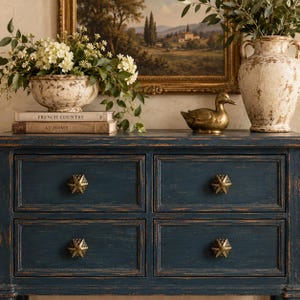 May include: A dark blue wooden dresser with four drawers featuring distressed edges and hexagonal brass knobs. On top sit two weathered cream-colored ceramic vessels, a brass duck figurine, and two books titled French Country and At Home.