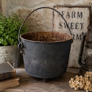 May include: A dark gray, round cast iron pot with a weathered, rusted interior and a thin, arched metal handle. The pot features three small, sturdy feet at the base and a textured, matte exterior finish, showcasing a heavy, industrial appearance.