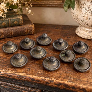 May include: A collection of ten round metal buttons featuring a dark, weathered finish with a textured, rope-like border and a central raised loop for attachment. Each button has a distressed, aged appearance with subtle gold-toned highlights.