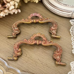 May include: Two ornate, antique-style drawer pulls in a warm, metallic finish. The pulls feature a curved design with intricate detailing and a blend of gold and rose-gold tones. They are set against a wooden surface.