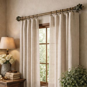 May include: Beige linen curtains hanging from a decorative gold-toned rod with ornate finials. The curtains frame a wooden window with a view of greenery. A lamp and side table with books and flowers are in the foreground.