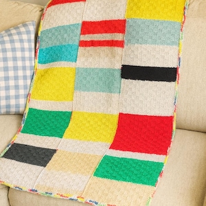 May include: A colorful, patchwork-style knitted blanket with a variety of rectangular blocks in shades of red, yellow, green, blue, and gray. The blanket is draped over a beige sofa, showcasing its vibrant design and textured surface.