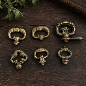 May include: Six antique brass drawer pulls in various designs. The pulls have a vintage aesthetic, with ornate details and a warm, aged finish. They are arranged on a dark wooden surface, showcasing their intricate shapes and textures.