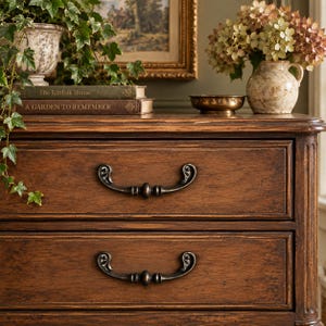 May include: A dark brown wooden dresser with two drawers featuring ornate metal handles. The top surface holds two stacked books titled The Kinfolk Home and A Garden to Remember, a potted ivy plant, a small brass bowl, and a ceramic vase with flowers.