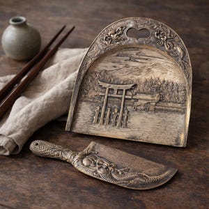 May include: A bronze-colored serving set featuring a tray with a landscape scene and a matching knife. The tray has a handle with dragon detailing. The knife also has dragon detailing on the handle and blade. The set is displayed on a wooden surface.