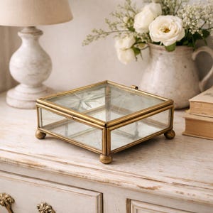 May include: A square, glass-topped jewelry box with a gold-colored metal frame. The box has a hinged lid and rests on four small, rounded feet. The glass top features a starburst design. A decorative item for storing jewelry.