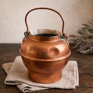 May include: A small, antique copper pot with a curved handle. The pot has a rounded body and sits on a tan linen cloth. The copper has a warm, reddish-brown hue, and the handle is attached with decorative loops.
