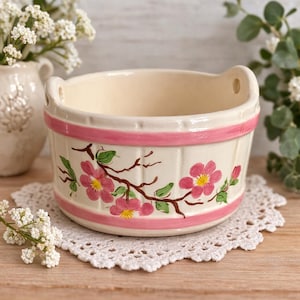 May include: Cream-colored ceramic bowl with pink floral design and handles. The bowl features a pink band around the top and bottom, with a floral pattern of pink flowers, green leaves, and brown branches. The bowl is sitting on a white lace doily.