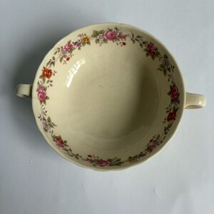 May include: Cream-colored ceramic soup bowl with two handles. The rim is decorated with a floral pattern of pink, red, yellow, and purple roses with green leaves. The bowl has a slightly scalloped edge.