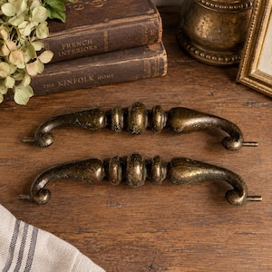 May include: Two matching drawer pulls with a dark, weathered bronze finish. Each handle features a central rounded bead flanked by smaller rings, tapering toward curved ends with small mounting posts extending from the outer edges.