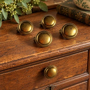 May include: A set of five round cabinet knobs with a distressed antique brass finish. Each knob features a smooth, domed surface and a circular base, designed for attachment to furniture drawers or doors, showcasing a weathered metallic texture.