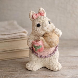 May include: A cream-colored bunny figurine with a pink bow, holding a teapot and a pink and red container. The bunny wears a pink apron with a purple trim. The figurine is on a wooden surface.