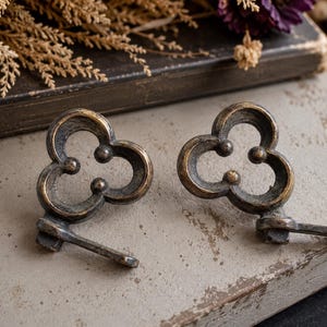 May include: Two antique bronze-colored key-shaped cabinet pulls. Each pull features a clover-like design with three rounded lobes and a small, rectangular key stem. The pulls are set against a neutral, textured background.