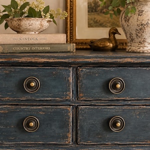 May include: A dark blue wooden dresser with a distressed finish and four drawers, each featuring a circular dark metal knob with a brass center. The dresser surface holds two stacked books titled The Kinfolk Home and Country Interiors.