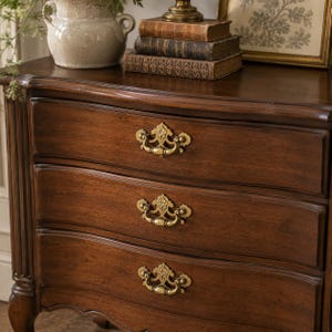 May include: A dark brown wooden chest of drawers featuring three horizontal drawers, each equipped with ornate brass swing handles. The piece has curved drawer fronts, fluted side columns, and cabriole legs, showcasing a polished wood grain finish.