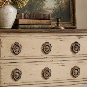 May include: Cream-colored antique dresser with six ornate bronze drawer pulls. The distressed finish reveals hints of the wood beneath. A vase of dried flowers, books, and a candlestick sit atop the dresser, with a framed landscape painting in the background.