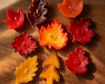 May include: A collection of leaf-shaped candles in various autumnal colors, including red, orange, and yellow. Each candle has a wick and is arranged on a wooden surface, creating a warm, seasonal display.
