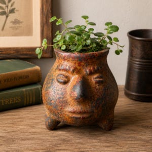 May include: A round, three-legged ceramic planter with a weathered, mottled orange and brown finish. The planter features a molded human face with closed eyes, a prominent nose, and a neutral mouth. A small green leafy plant grows inside.