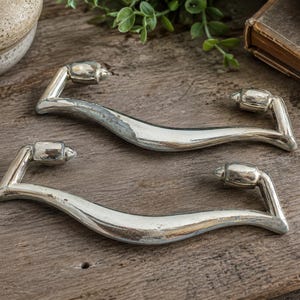 May include: Two silver-toned cabinet pulls with a curved design. Each pull has a central, wave-like handle and angled end pieces with rounded, decorative accents. The pulls are displayed on a weathered wooden surface, suggesting a vintage or rustic aesthetic.