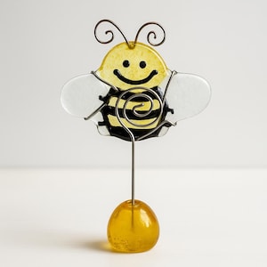May include: A whimsical bee-shaped photo holder. The bee features a yellow body with black stripes, clear wings, and a smiling face. The bee is made of glass and wire, and sits atop a yellow glass base. The antennae are made of wire.