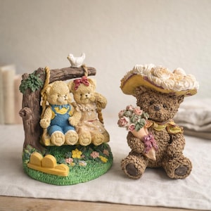 May include: Two teddy bear figurines. One features two bears on a swing, with a small bird perched above. The other bear is brown, wearing a hat and holding a bouquet of flowers. The base is green with flowers.