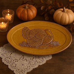May include: Oval, mustard-colored serving platter with a detailed relief of a turkey. Two small pumpkins and two lit votive candles are in the background. A white lace doily is in the foreground. Thanksgiving-themed tableware.